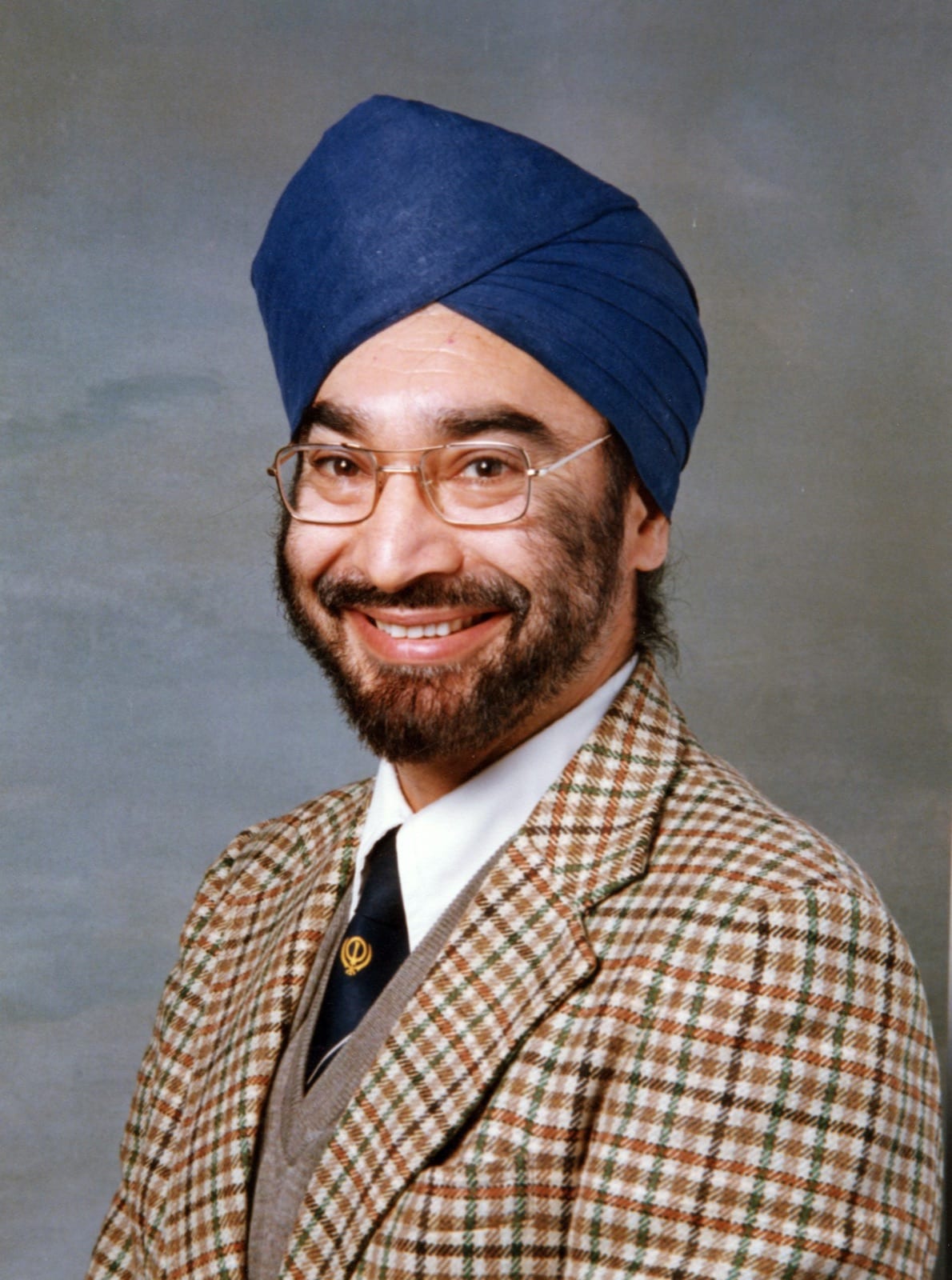 Portrait of Avtar Virdee, smiling warmly in a blue turban and glasses, wearing a checked tweed jacket and a tie pinned with a khanda.