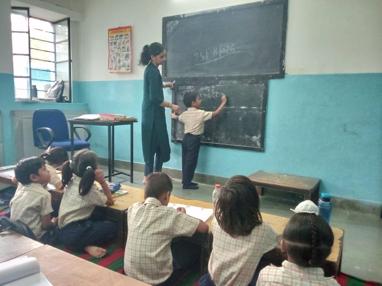 A young boy practises writing letters on the blackboard while his teacher stands beside him, classmates watching from their desks.