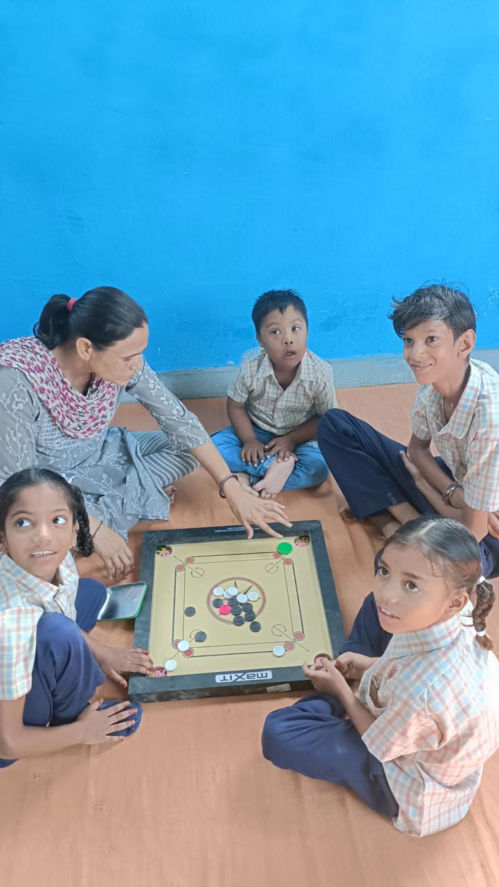 A teacher and four children sit on a mat around a carrom board against a bright blue wall, one child reaching to take her shot.