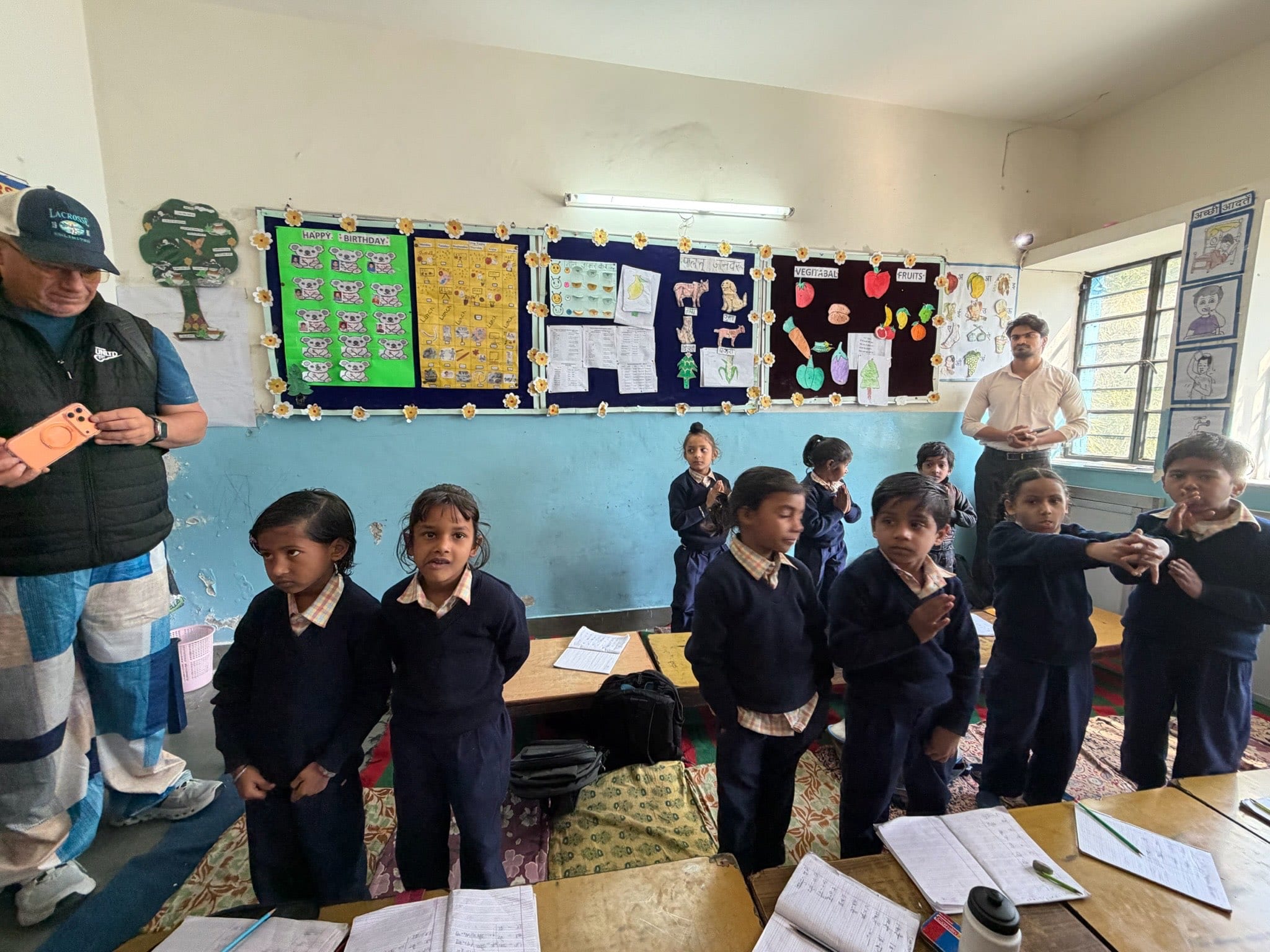 A visitor photographs the class on his phone while pupils stand by a wall of birthday charts and cut-out decorations.