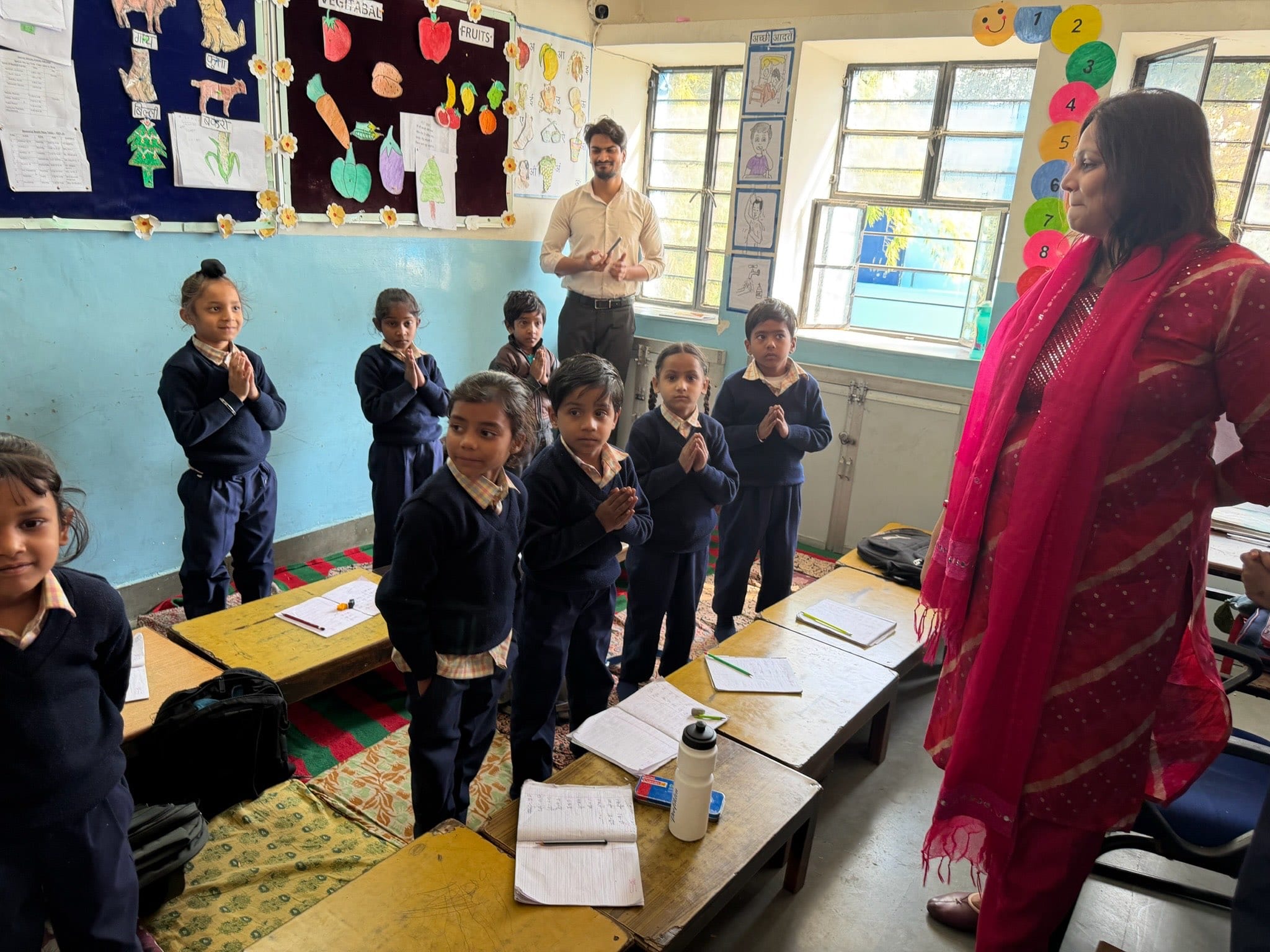 Pupils stand at their desks with hands pressed together in welcome while two teachers stand at the back of the sunlit classroom.