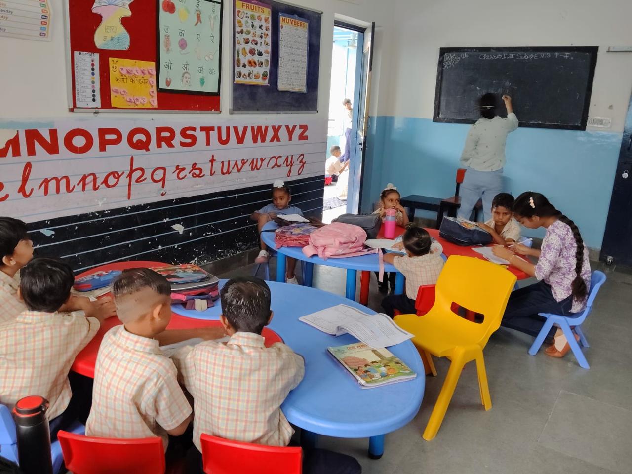 Infant classes work at bright blue and red tables beneath a hand-painted alphabet wall while a teacher writes on the blackboard.