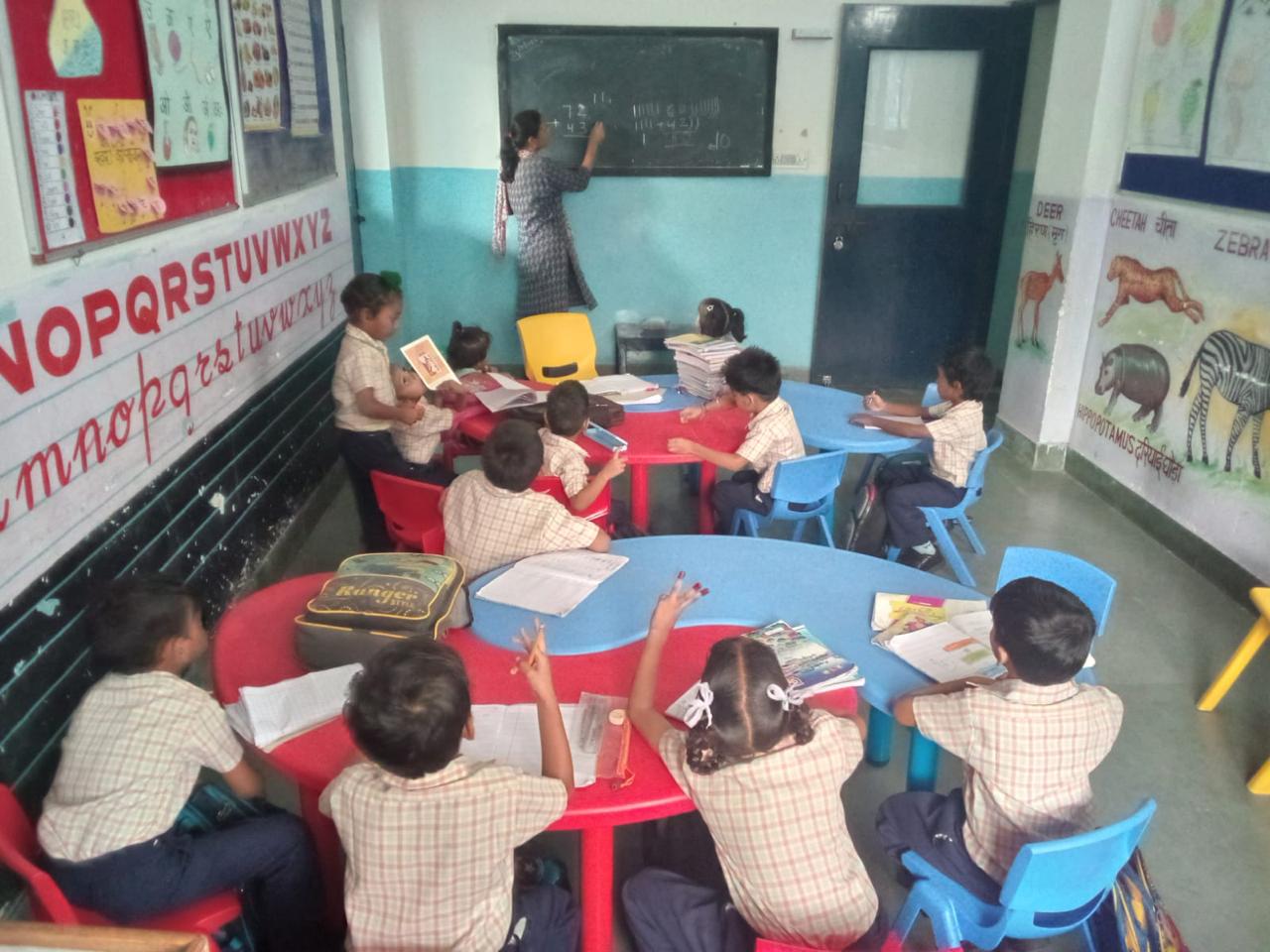 A teacher writes sums on the blackboard while young children work at curved red and blue tables, the walls painted with alphabets and animals.