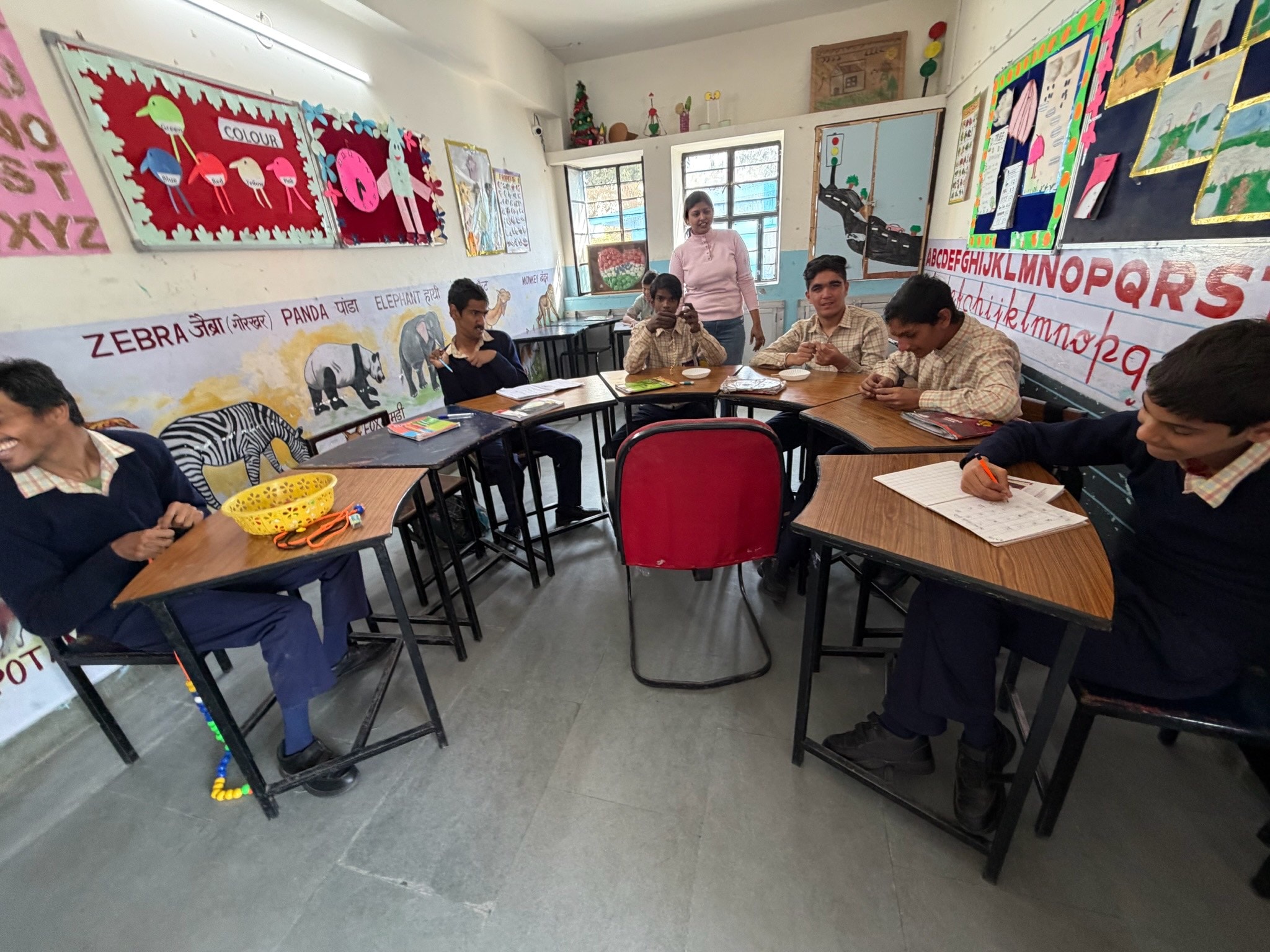 Students share a joke during a lesson at the Prayas school; a boy in the foreground writes in his exercise book while a classmate across the curved desks laughs, the walls painted with animals and the alphabet.