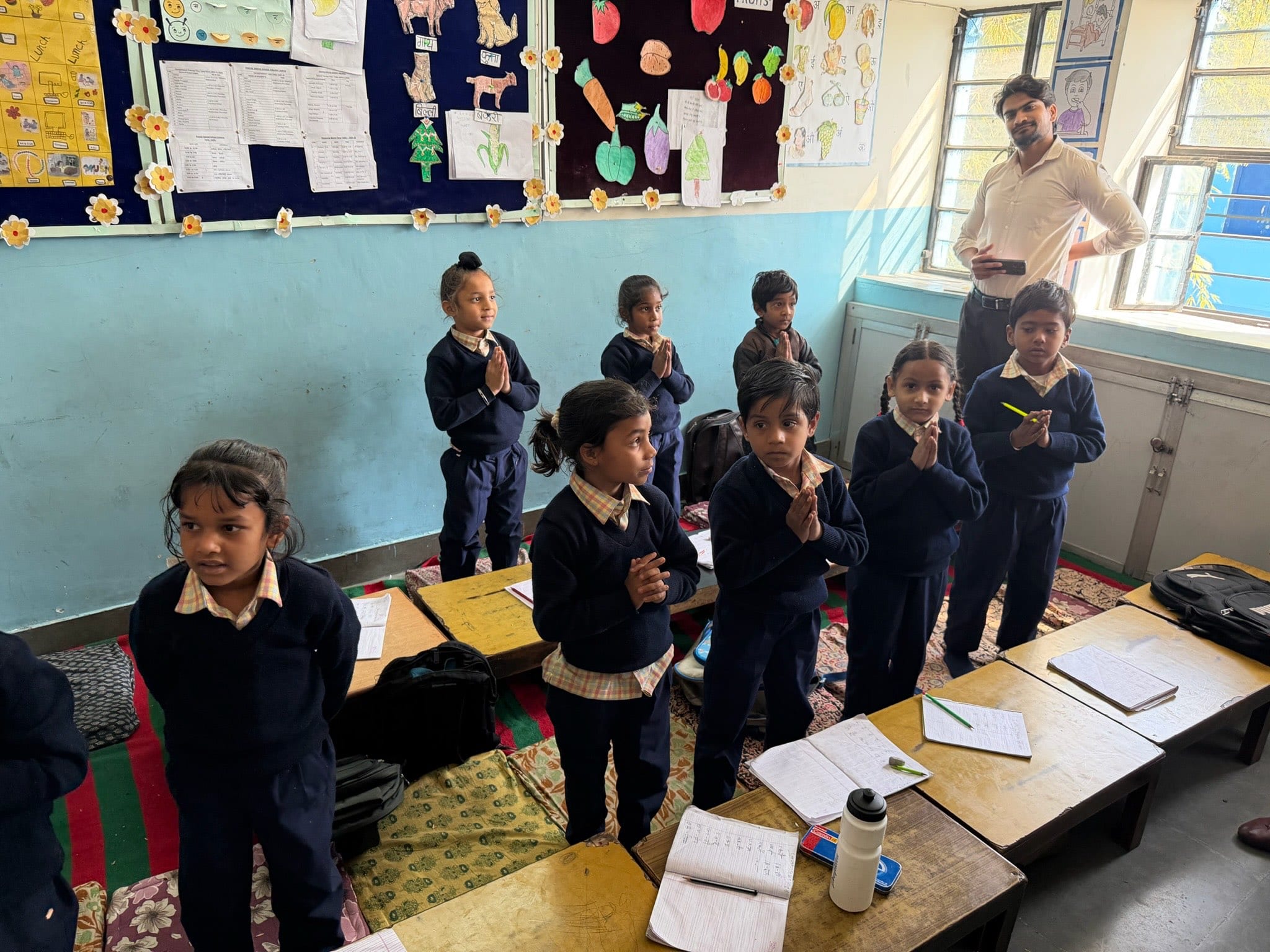 Young children in navy school jumpers stand by their low desks with hands pressed together in greeting, notebooks open in front of them.