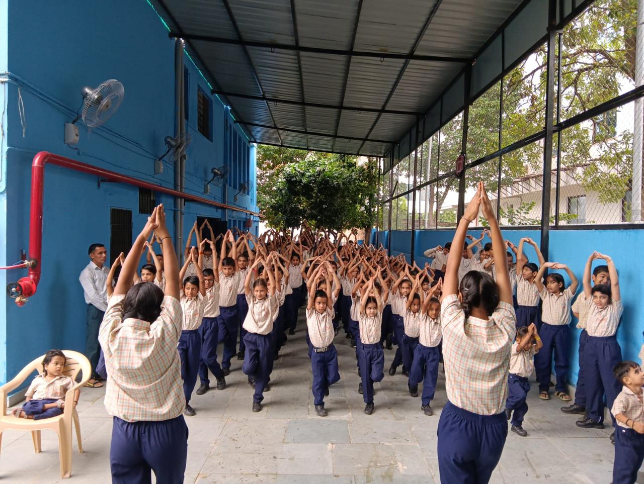 The whole school practises yoga together in the covered courtyard, arms raised overhead, with two older pupils leading from the front.