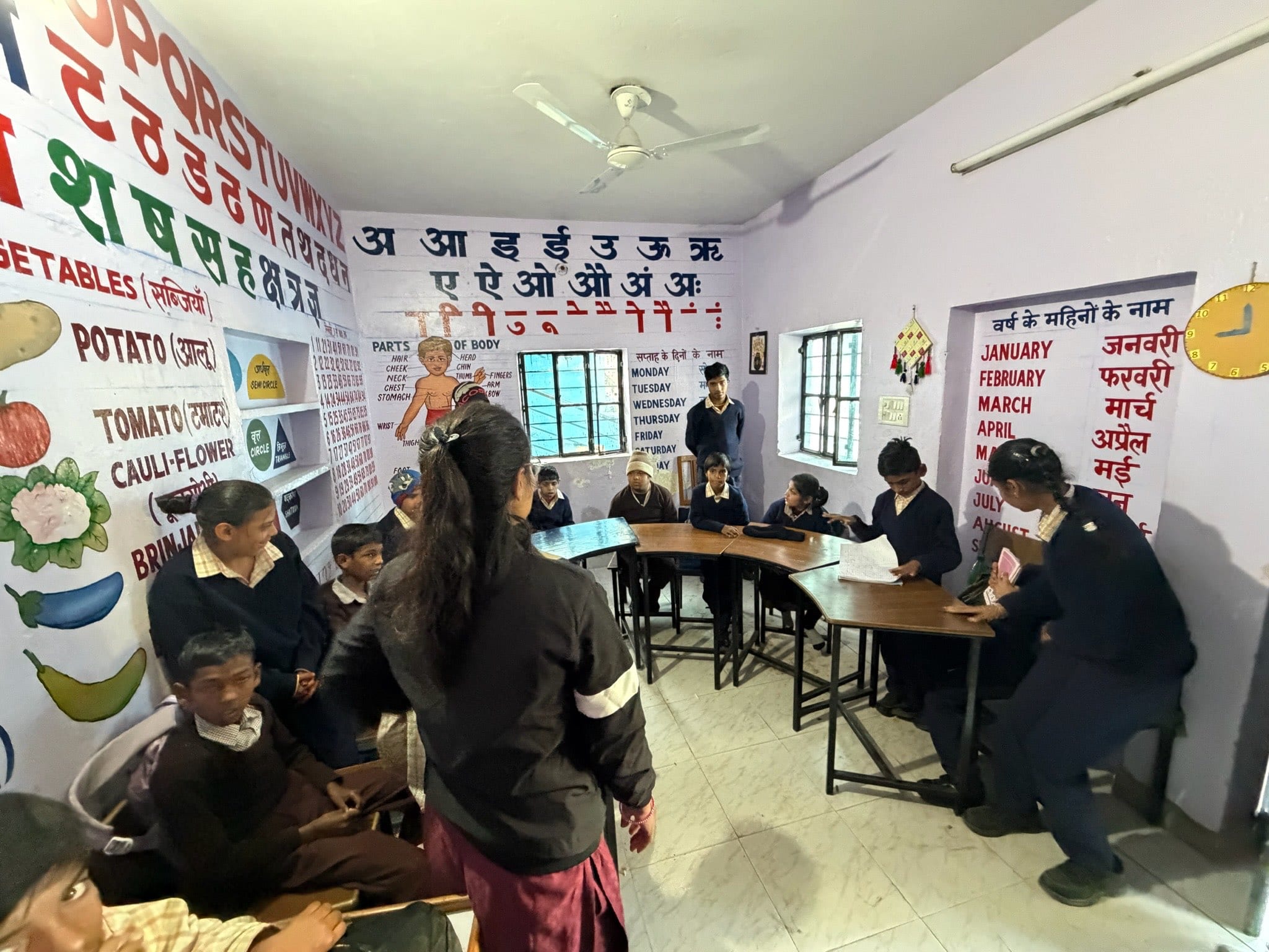 A classroom whose walls are painted edge to edge with alphabets, vegetables and the months of the year; students sit around curved tables while one reads aloud from her notebook.