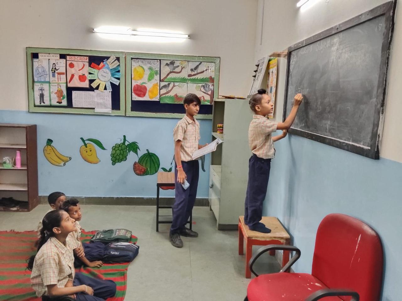 Two pupils write on the classroom blackboard - one standing on a stool to reach - while younger children watch from a mat, fruit murals along the wall.