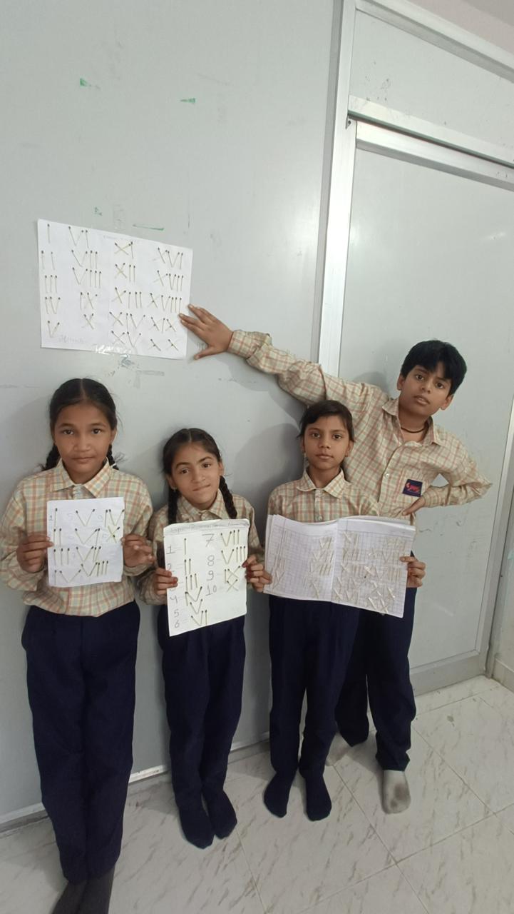 Four pupils hold up Roman numerals they have made from matchsticks; one reaches up to point at a finished sheet pinned to the wall.