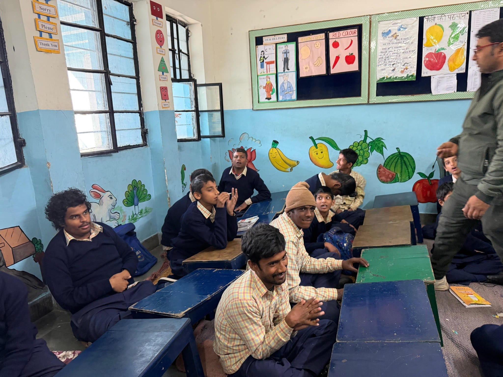 Older students sit at low blue and green desks in a classroom painted with fruit murals; several look up smiling as the photo is taken.