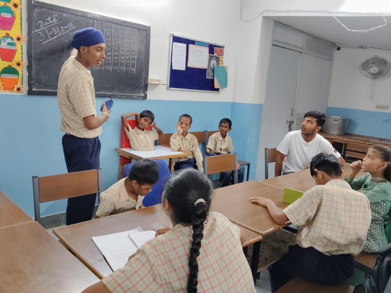 A pupil stands at the blackboard leading the class through the times tables he has chalked up, watched by his classmates and a teacher.