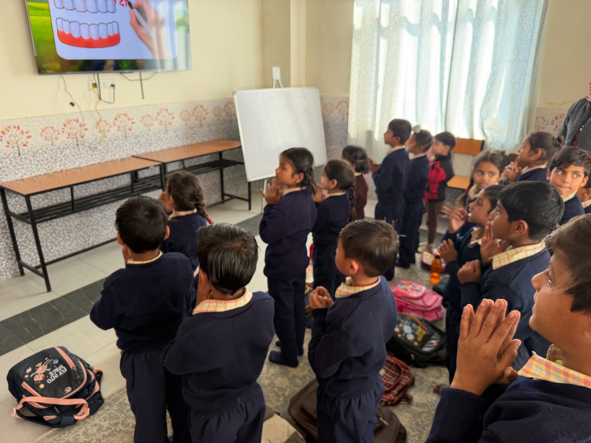 Children stand together with hands folded, watching a lesson playing on a wall-mounted screen at the front of the room.